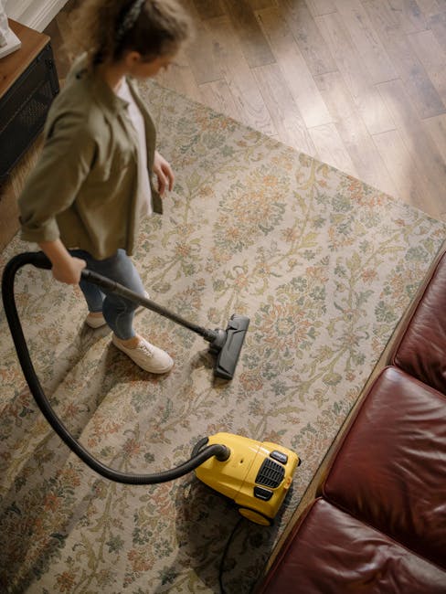 An interior living room scene showing a wooden sideboard with a white bust sculpture, books, and a remote control on top. The room features a patterned area rug on a wooden floor, with a yellow vacuum