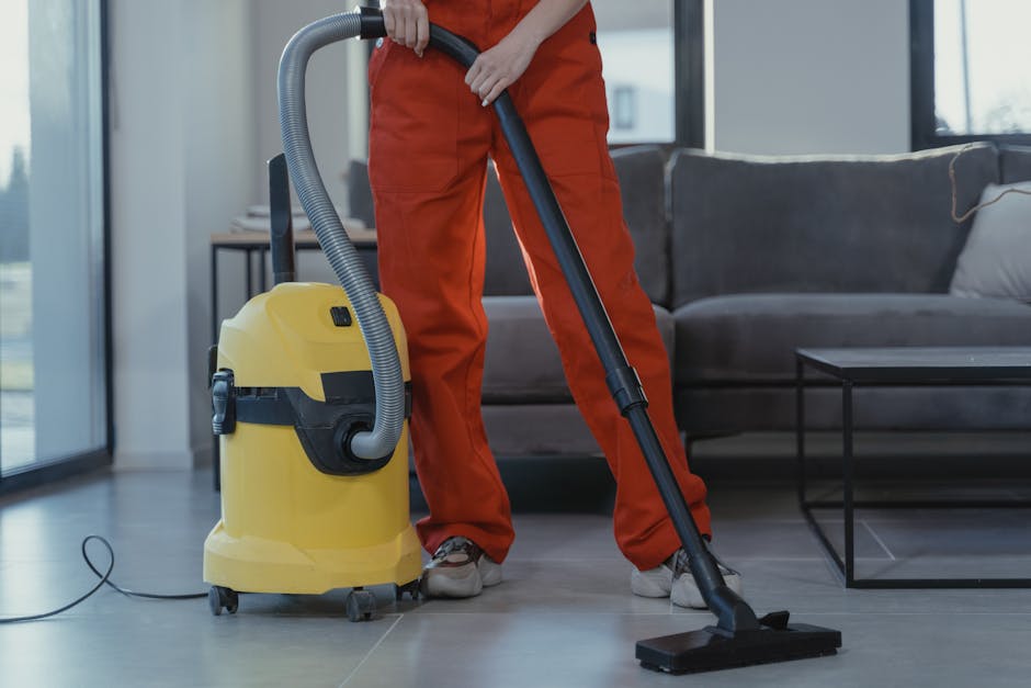 A person dressed in orange cleaning overalls using a yellow vacuum cleaner with a black hose attachment on a light-colored tiled floor in a modern living room. The room features large windows allowing natural light, with a gray fabric sofa and a black glass coffee table visible in the background. The surfaces are clean, with no dust or debris, and the vacuum cleaner is actively engaged in surface cleaning. Chelsea Carpet Cleaners provide professional domestic cleaning and deep carpet sanitisation services, ensuring a high standard of hygiene in residential spaces.