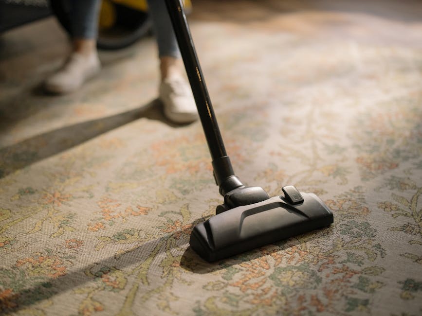 A close-up of a vacuum cleaner head being used to deep clean a patterned, ornate carpet in a residential living room. The carpet features intricate floral motifs in beige, green, and orange tones. The vacuum attachment is black with a visible switch, and a person's white sneakers and the lower part of their trousers can be seen in the background. Natural light illuminates the scene, highlighting the cleanliness and freshness of the carpet. This image demonstrates surface cleaning and maintenance as part of a professional domestic cleaning service provided by Chelsea Carpet Cleaners, emphasizing thorough sanitisation of home textiles.