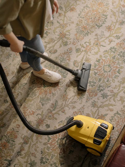 A person dressed in orange cleaning overalls using a yellow vacuum cleaner with a black hose attachment on a light-colored tiled floor in a modern living room. The room features large windows allowing natural light, with a gray fabric sofa and a black glass coffee table visible in the background. The surfaces are clean, with no dust or debris, and the vacuum cleaner is actively engaged in surface cleaning. Chelsea Carpet Cleaners provide professional domestic cleaning and deep carpet sanitisation services, ensuring a high standard of hygiene in residential spaces.