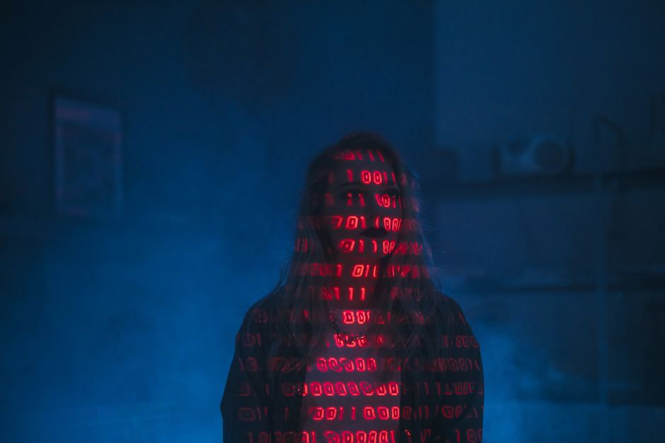 A person with long hair standing in a dimly lit room, with their face and upper body illuminated by red digital code projectors displaying binary numbers across their face and clothing. The background